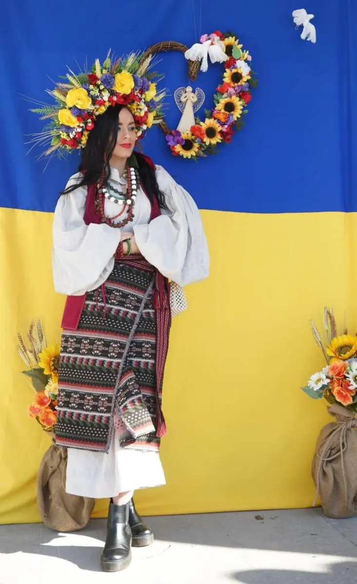 A woman in an elaborate Ukrainian folk costume and flower crown standing in front of a large Ukraine flag backdrop at UAFest