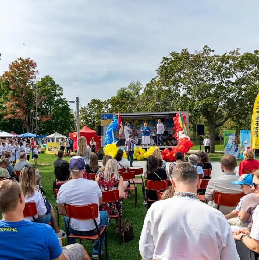 Wide shot of a festival stage performance framed by balloon decor, with a large crowd seated in lawn chairs on the grass