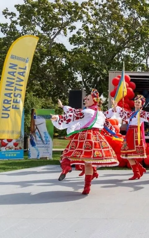 Two female folk dancers mid-performance on stage in red, white and gold costumes with raised arms