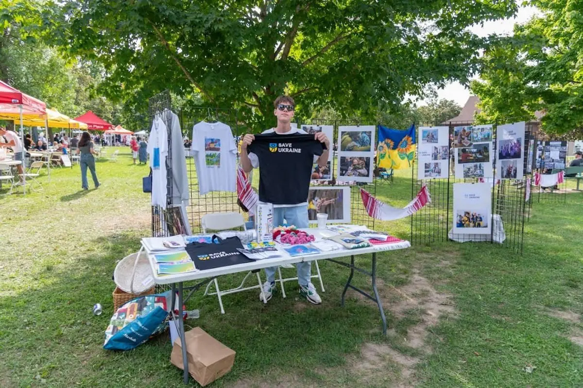 Cultural exhibit tent with an embroidered towel and folk-art photo displays on wire boards, sunflower artwork, festival-goers walking through the park behind