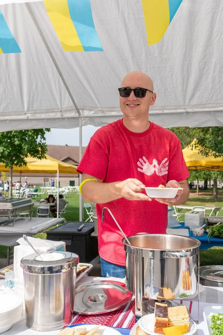 A volunteer in a Ukrainian Fest t-shirt serving a plate of food from a large pot under the festival food tent