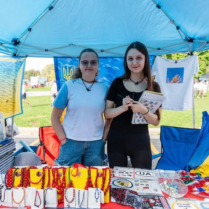 Two young women staffing a craft stall table filled with colorful handmade beaded jewelry and Ukrainian-themed souvenirs