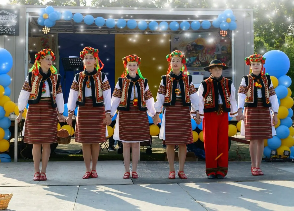 Six children and teens in traditional Ukrainian folk costume holding hands on stage in front of a balloon arch
