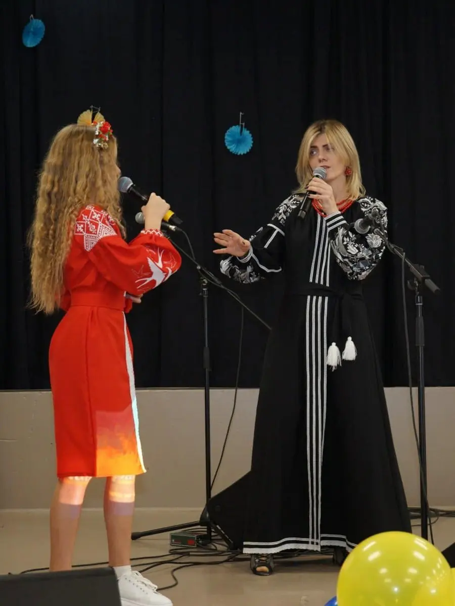 A teenage girl in a flower crown and red embroidered dress singing into a microphone on stage beside a host