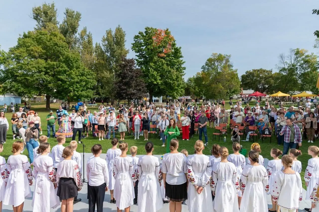 Crowd of visitors gathered on the grass at Riverside Park watching a group of performers in white embroidered Ukrainian shirts at UAFest