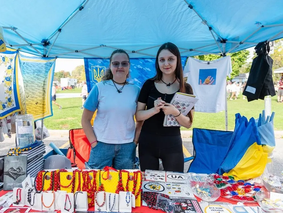 A vendor holding up a 'Save Ukraine' t-shirt at a UAFest merchandise table, with flags and support materials laid out for visitors