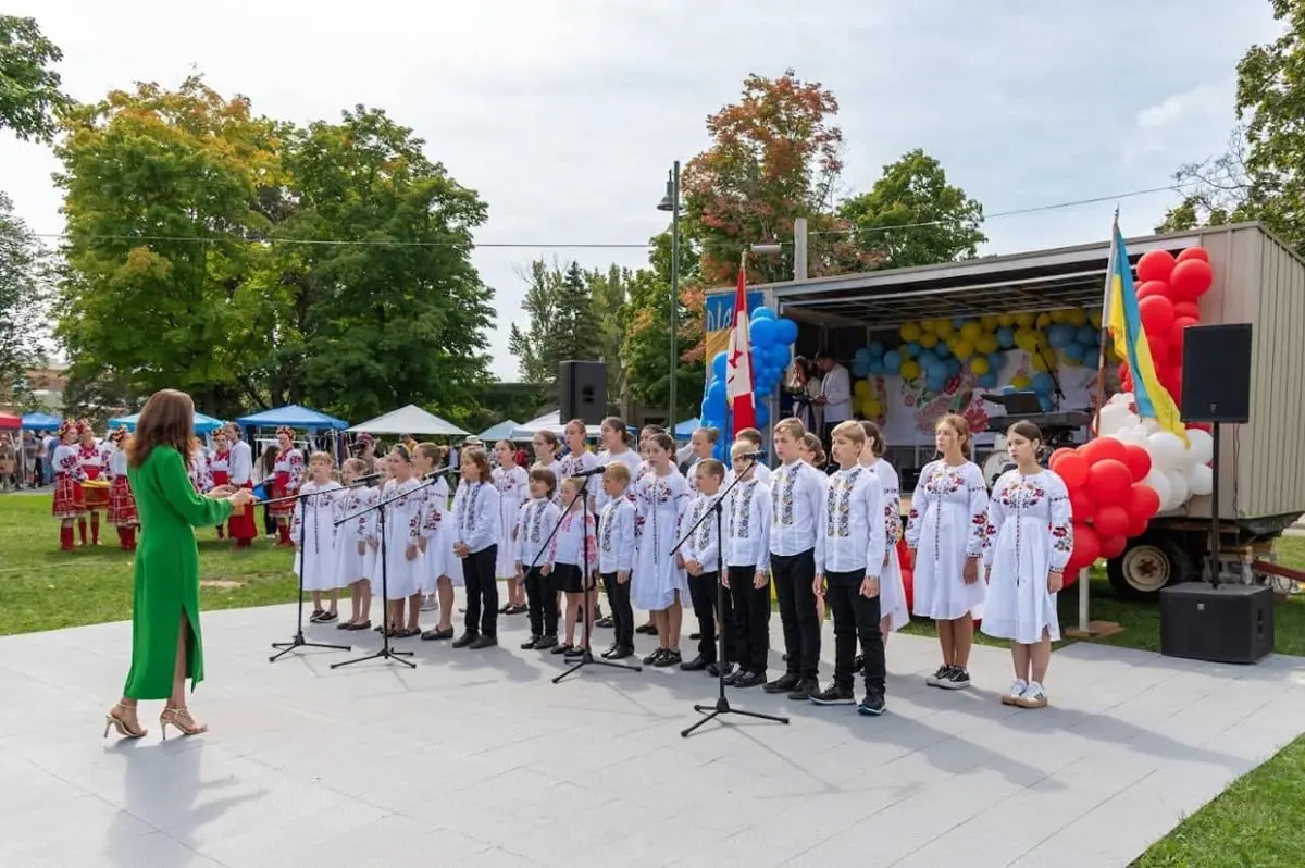 Children and youth in white Ukrainian embroidered vyshyvanka shirts standing in a circle on the festival lawn, facing a large crowd of guests