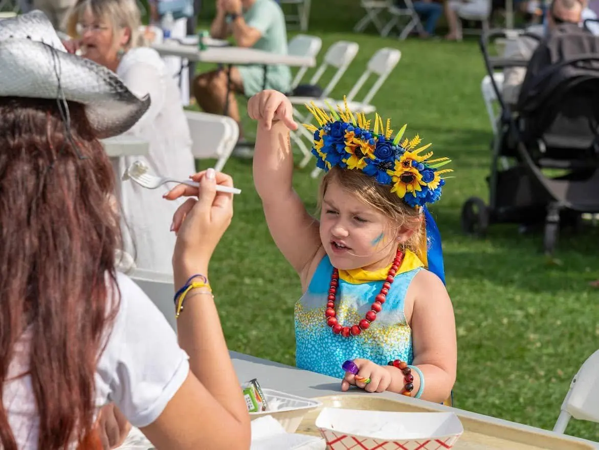 A young girl in a blue-and-yellow sunflower crown at a picnic table in the festival kids' area