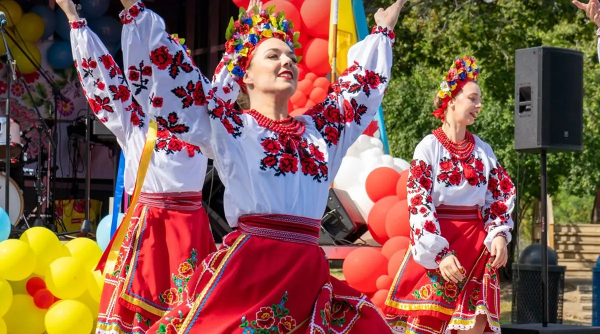 Folk dancers in red embroidered skirts, white embroidered blouses, and floral crowns dancing with arms raised on the UAFest stage