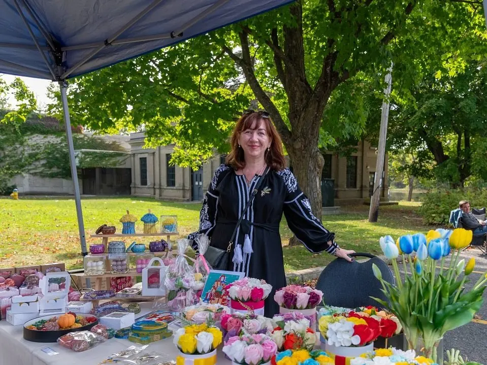 A vendor smiling behind a market tent table of handmade soaps, gift boxes and fresh flower bouquets at the UAFest Ukrainian Market