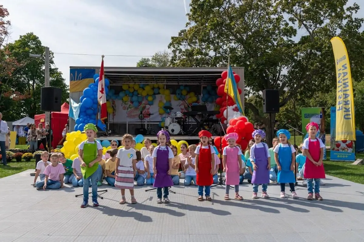 Children in colourful chef aprons and hats singing on the UAFest stage, framed by blue, yellow and red balloon decorations