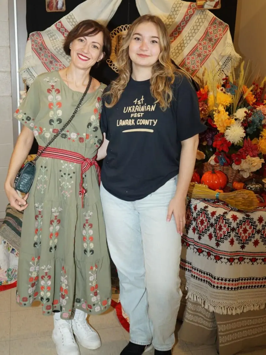A volunteer wearing a '1st Ukrainian Fest Lanark County' t-shirt stands with a woman in an embroidered vyshyvanka dress in front of a heritage textile and flower display.
