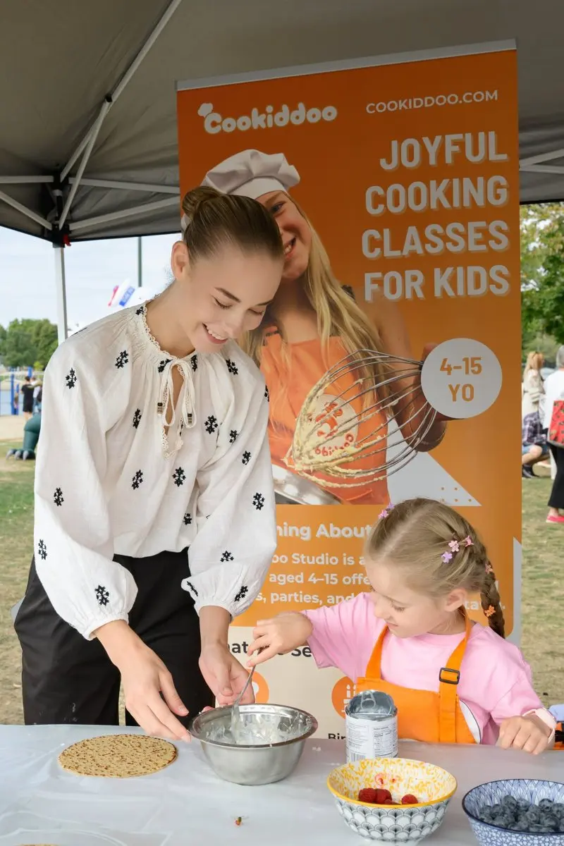 A teen volunteer in an embroidered blouse helps a young girl in a pink apron mix batter at the Cookiddoo booth in the Kids' Zone.