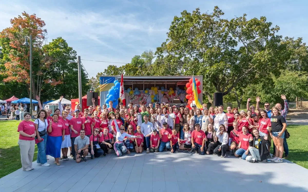 A large group of UAFest volunteers in matching red festival t-shirts posing together on the outdoor stage at Riverside Park during UAFest 2025.