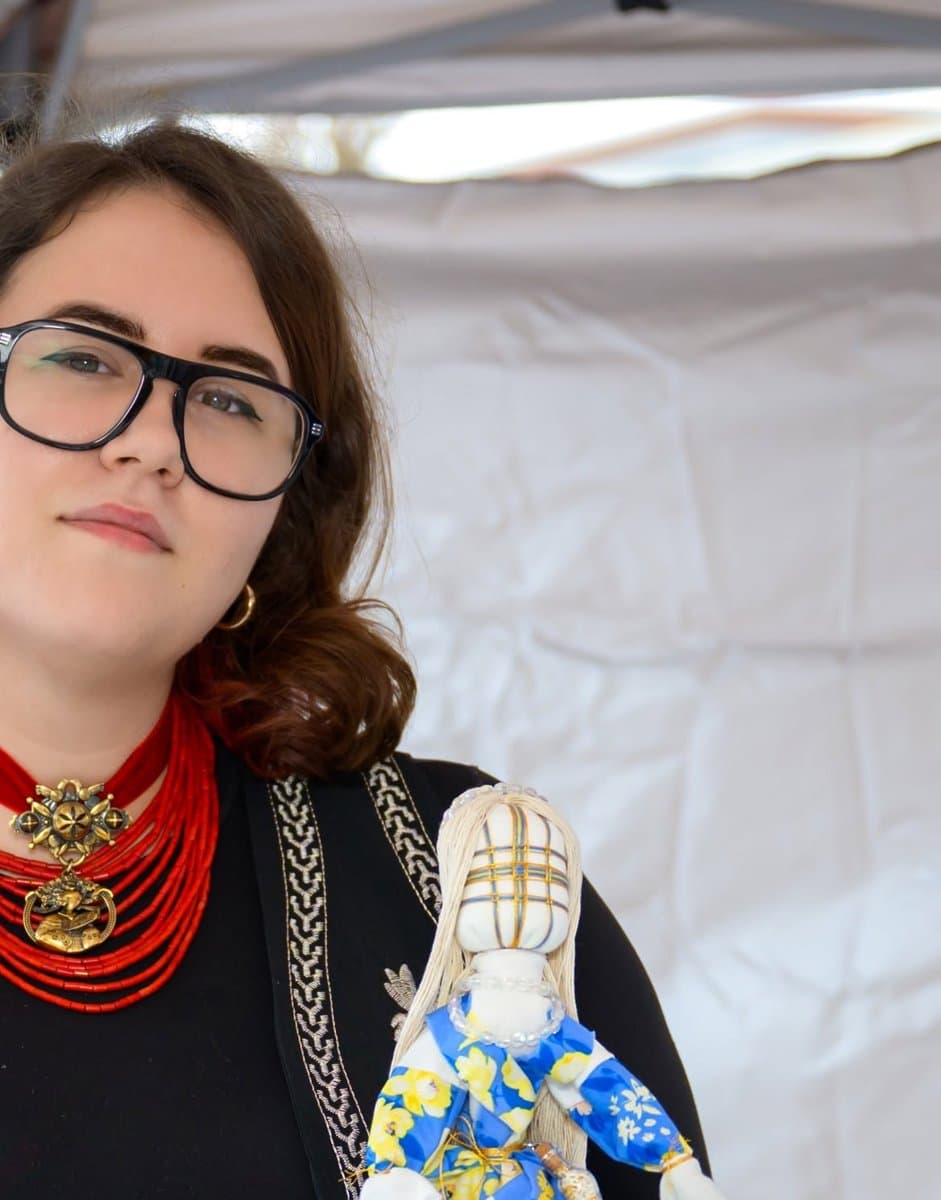 A community member wearing a red coral bead necklace holds a handmade Ukrainian motanka doll in a blue and yellow floral dress at UAFest