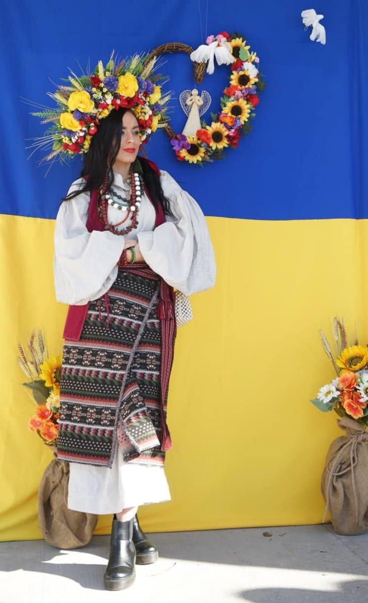 A woman in an elaborate Ukrainian folk costume and flower crown standing in front of a large Ukraine flag backdrop at UAFest