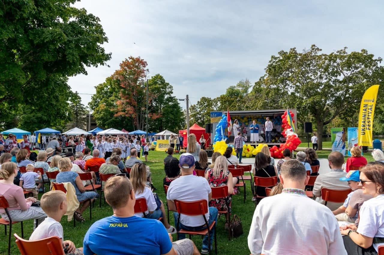A wide view of Riverside Park during the festival, showing the outdoor stage with balloon decorations, market tents, mature trees and the Mississippi River in the background