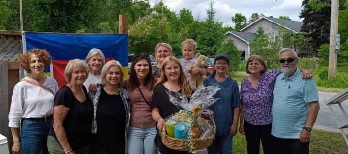 A group of UAFest organizers and supporters posing outdoors in front of a Ukrainian flag, holding a sunflower gift basket, at an early community fundraiser in Carleton Place