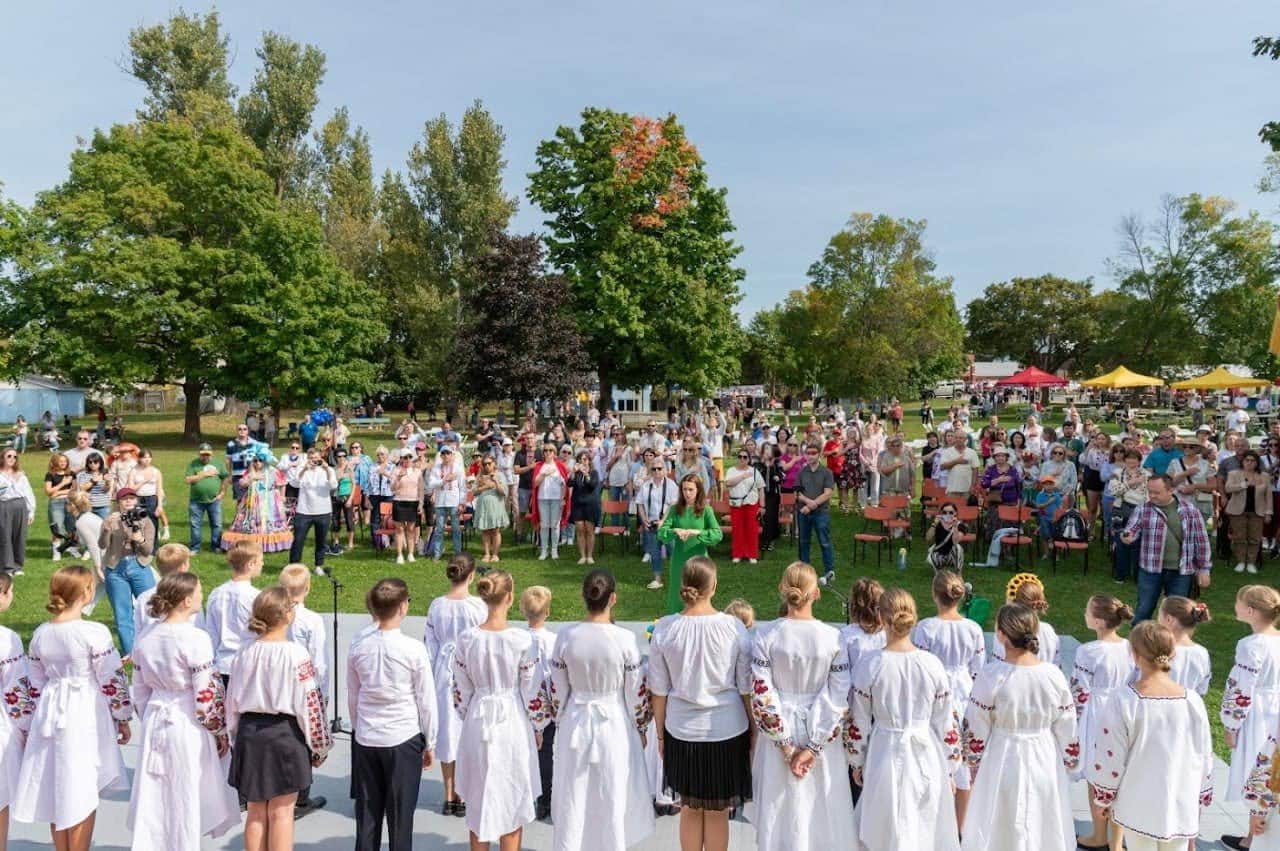 Crowd of visitors gathered on the grass at Riverside Park watching a group of performers in white embroidered Ukrainian shirts at UAFest