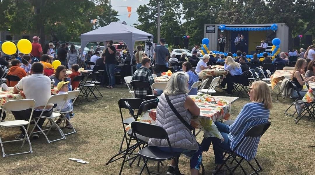 A wide crowd seated at tables with the stage and band in the background, balloon decorations overhead, at UAFest