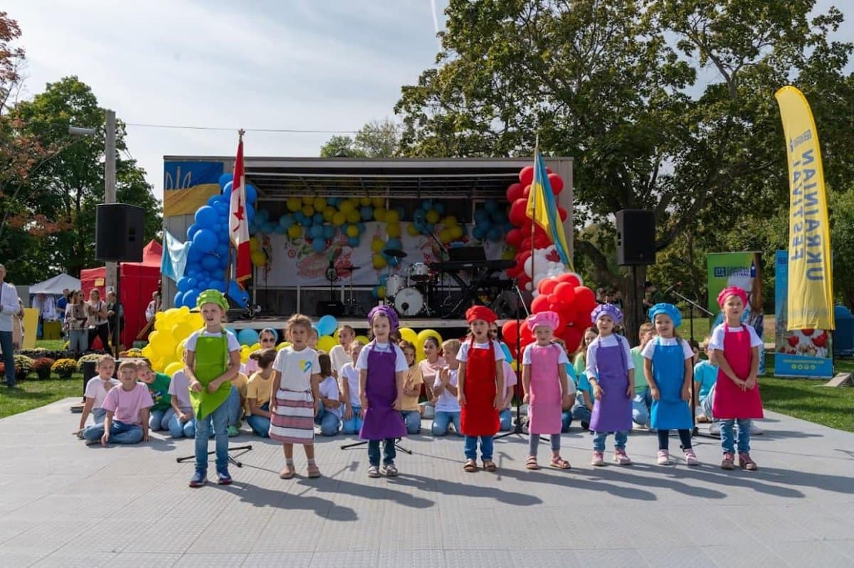Children in colorful chef aprons and hats singing on the UAFest stage, framed by blue, yellow, and red balloon decorations