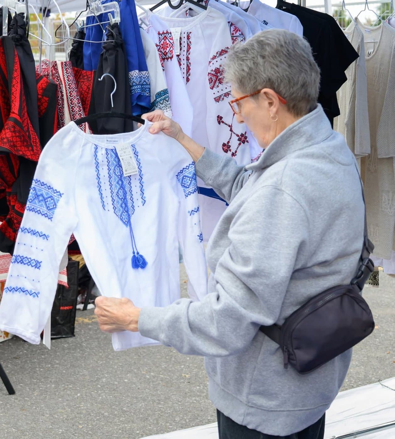 An elderly woman examining an embroidered white and blue vyshyvanka blouse hanging on a rack at a UAFest clothing vendor tent