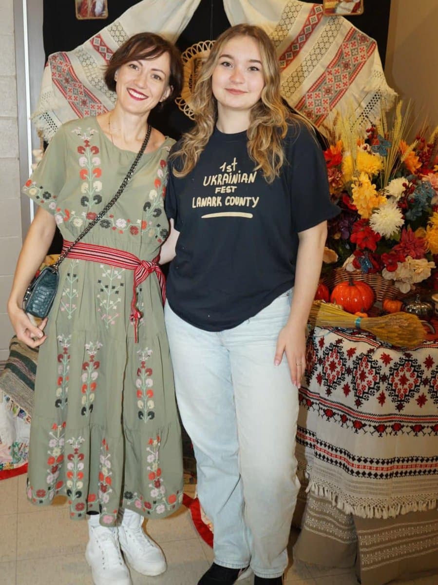 A volunteer wearing a '1st Ukrainian Fest Lanark County' t-shirt stands with a woman in an embroidered vyshyvanka dress in front of a heritage textile and flower display.