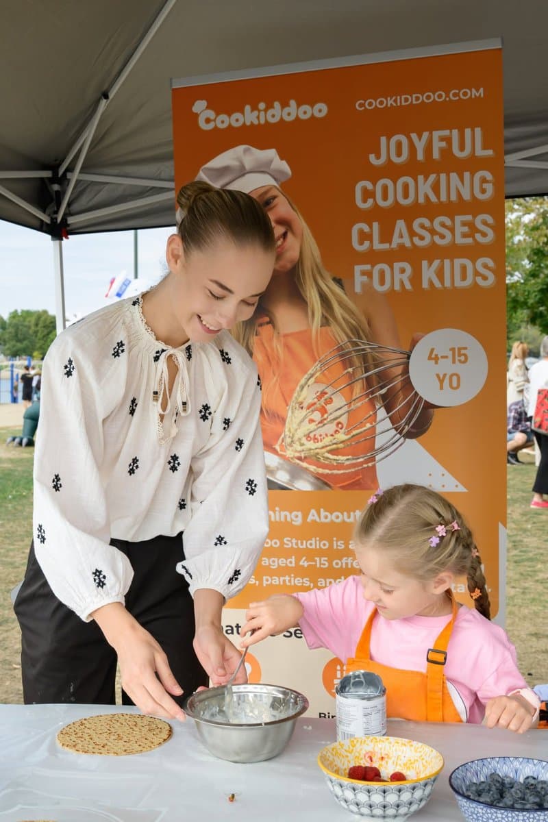 A teen volunteer in an embroidered blouse helps a young girl in a pink apron mix batter at the Cookiddoo booth in the Kids' Zone.