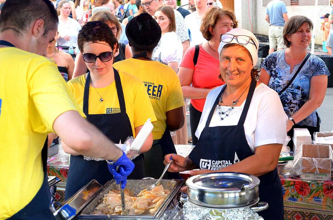 Festival volunteers in yellow shirts and Capital Ukrainian Festival aprons serve varenyky from a chafing tray to a queue of visitors