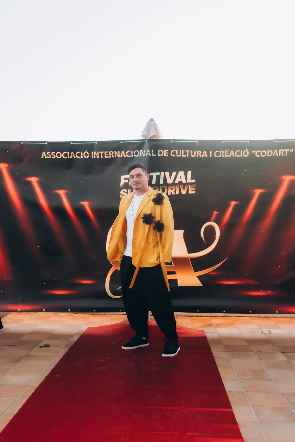 DEN BAURIN in a yellow jacket on a red carpet in front of the Festival Super Drive backdrop, Barcelona 2025