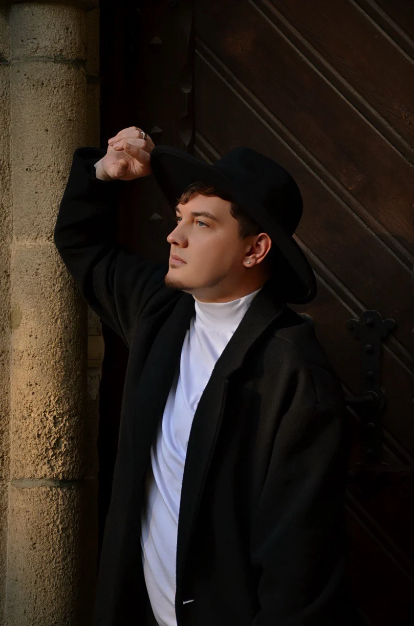 DEN BAURIN in a black coat and hat leaning against a stone column and dark wooden door, from his Autumn photoshoot gallery
