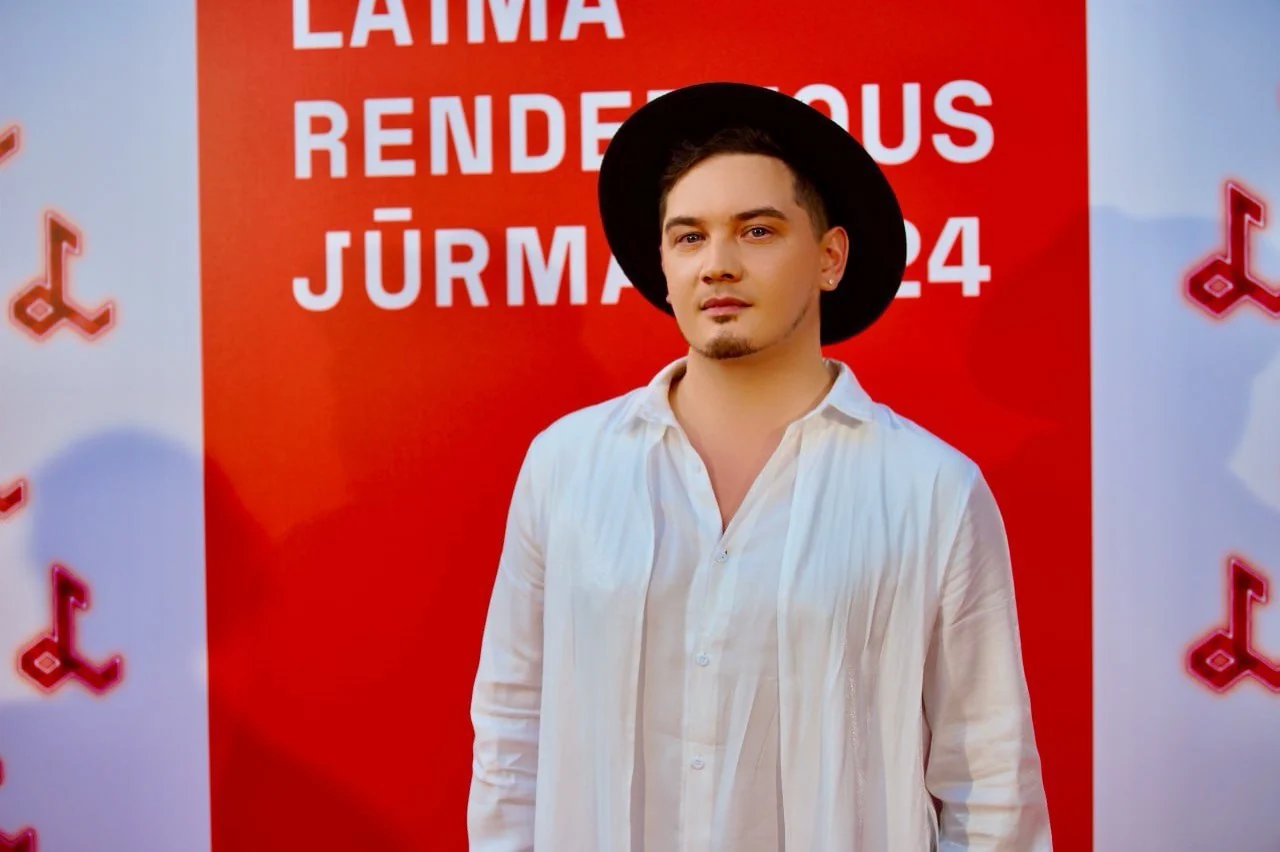 DEN BAURIN in a white shirt and black hat in front of the red Laima Rendezvous Jūrmala 2024 backdrop