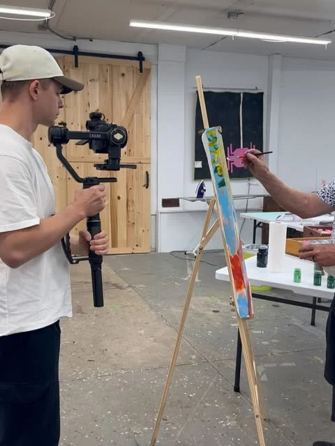 Denys filming on a gimbal in a bright workshop while, across the frame, a hand paints a tall canvas on an easel
