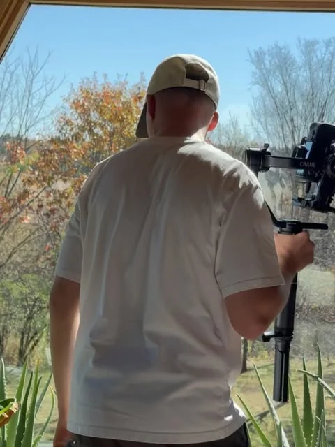 Denys seen from behind, camera on a gimbal, filming towards a window with autumn trees outside