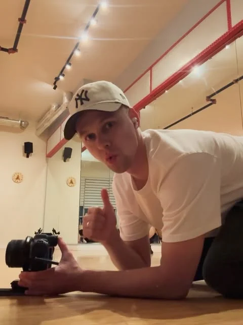 Denys lying on the floor of a mirrored dance studio with his camera at floor level, one finger raised to the lens