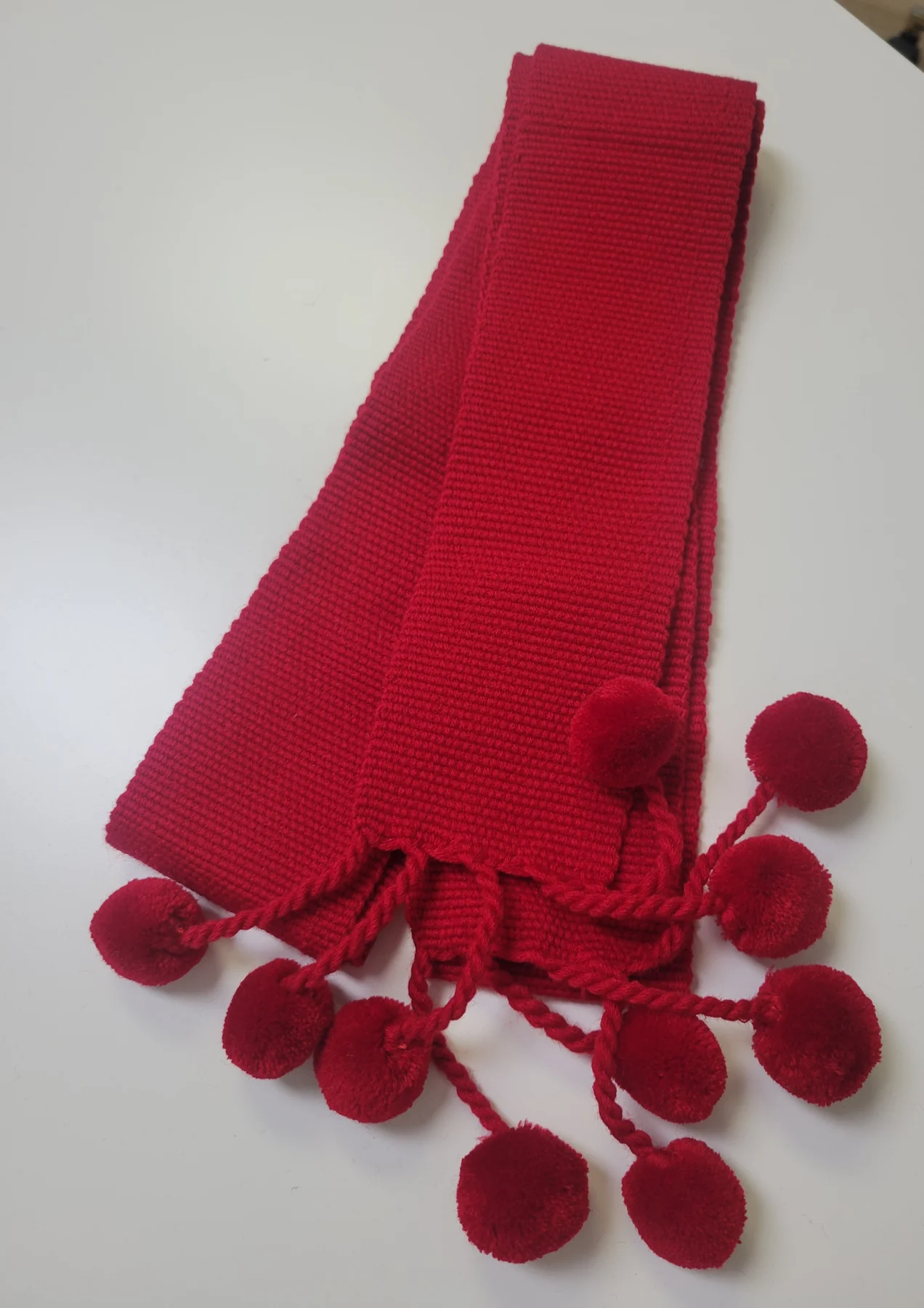 A red woven krayka, three metres long, finished with hand-made pom-poms