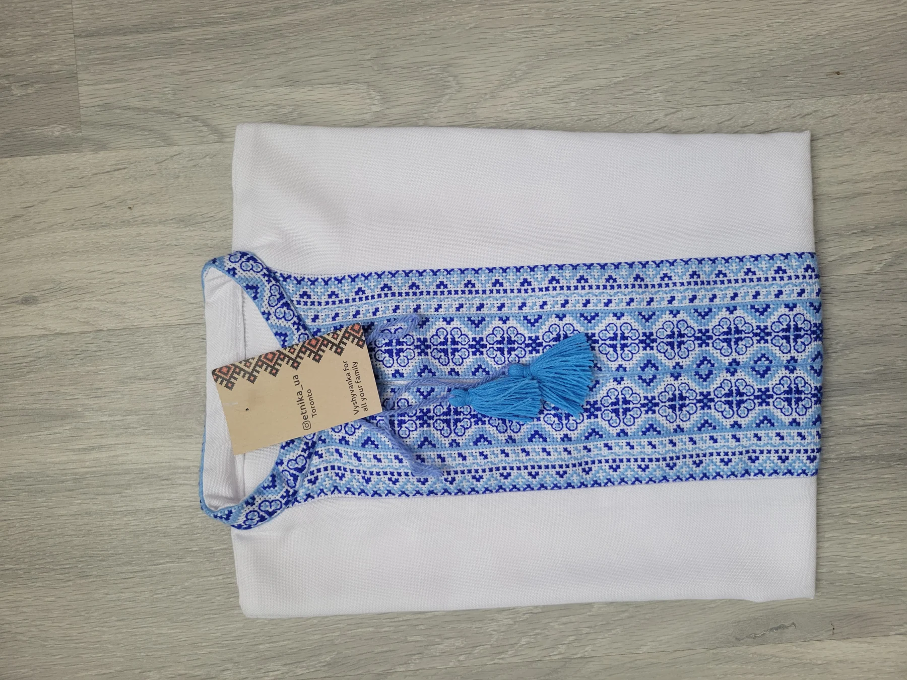 "Символ Волі" — a teenager's handmade vyshyvanka, white with blue embroidery and blue tassel ties, with the ETNIKA UA hangtag attached