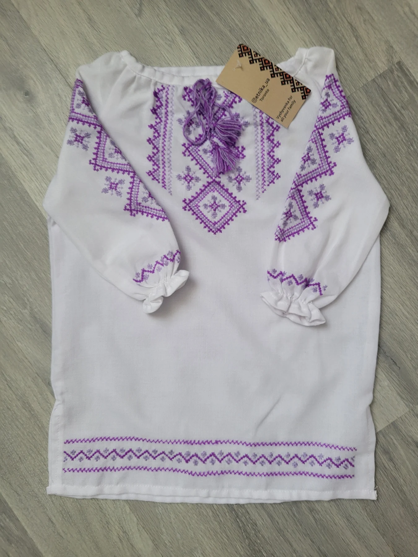 "Мальва" — a child's hand-embroidered shirt in violet thread, with the ETNIKA UA hangtag attached