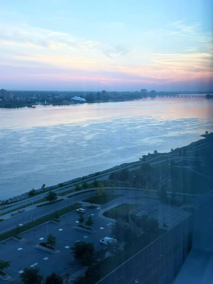 A wide river at dusk under a pink and blue sky, photographed from high up: a bridge and a low city skyline on the far bank, a riverside road and parking lot below