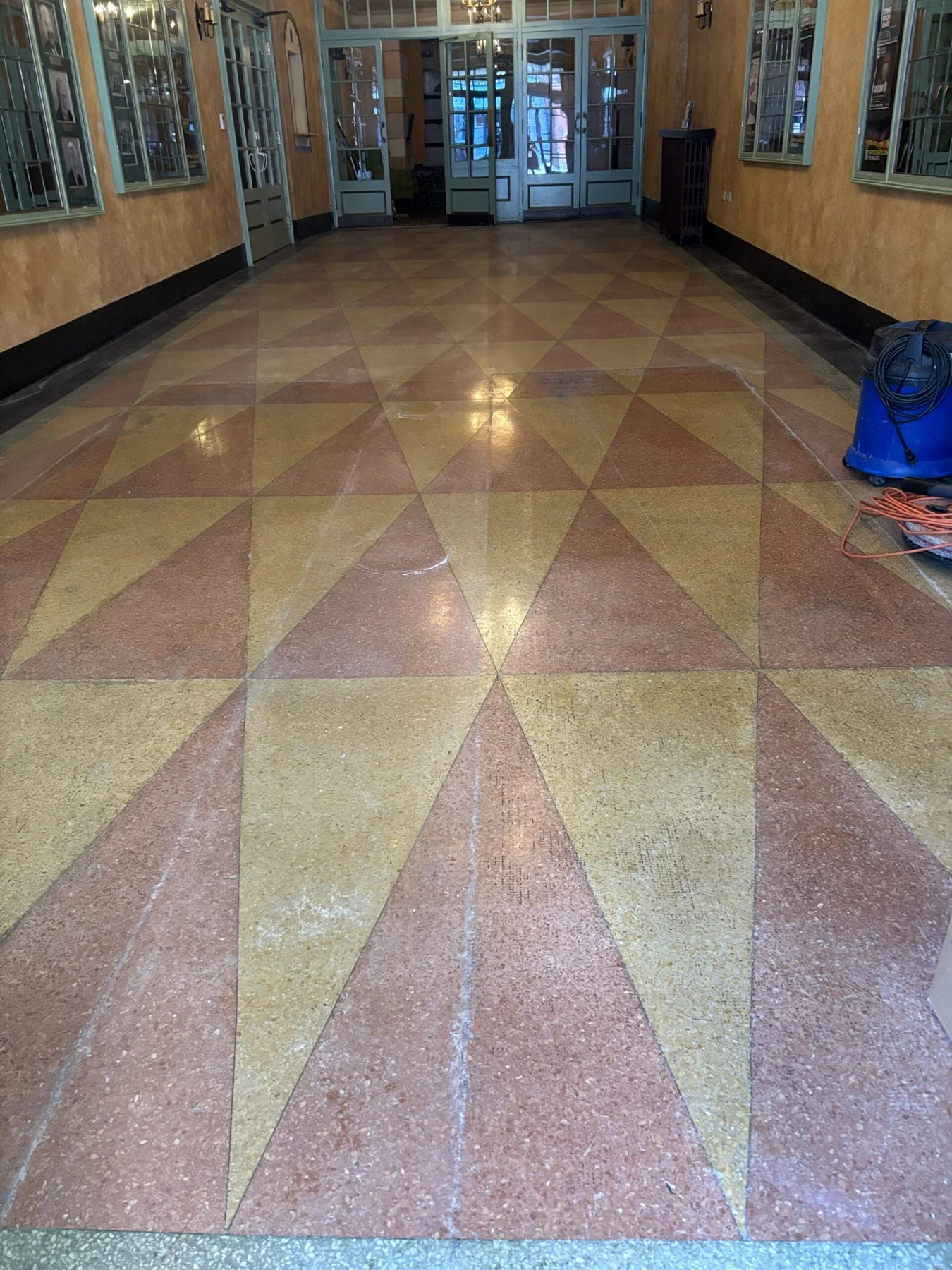 The Capitol Theatre entrance hall in Port Hope before the work: a dull, scuffed terrazzo floor in a red-and-gold triangular starburst pattern, with a blue wet vacuum and an orange extension cord at the right edge.