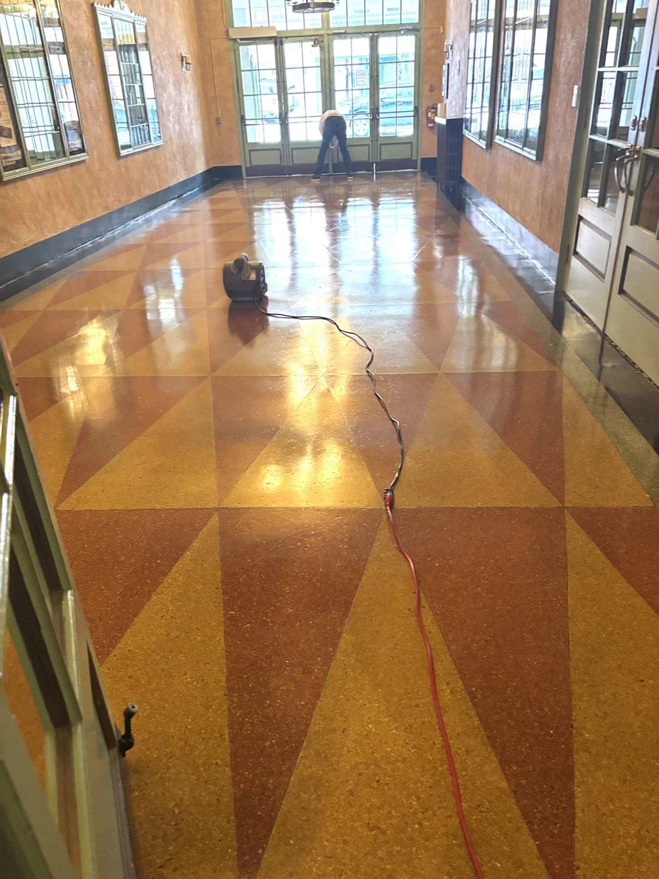 The same Capitol Theatre entrance hall after refinishing: the terrazzo now glossy enough to reflect the windows and doors, with a floor blower on the tiles and a worker bent over near the front doors.