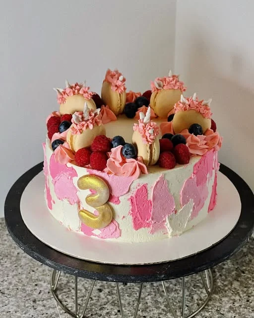 Birthday cake in pink and white buttercream with a gold number three, macarons, raspberries and blueberries