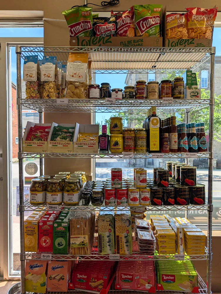 A window-side shelving unit of European pantry staples — pasta, pesto, olives, oils, hot sauces, crackers and Manner and Loacker wafers.