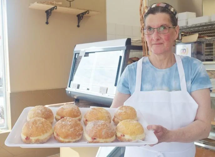Doris in an apron and headscarf holding a tray of freshly filled Berliners in front of the bakery display case.