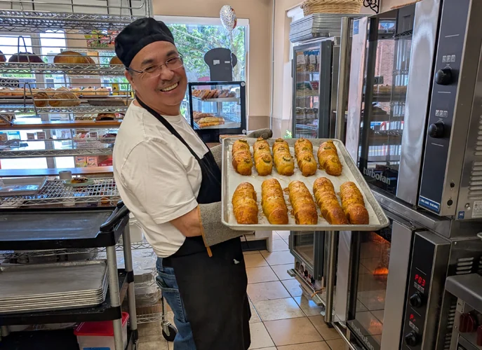 Rob in a baker's cap and apron carrying a sheet tray of fresh baking past the ovens.