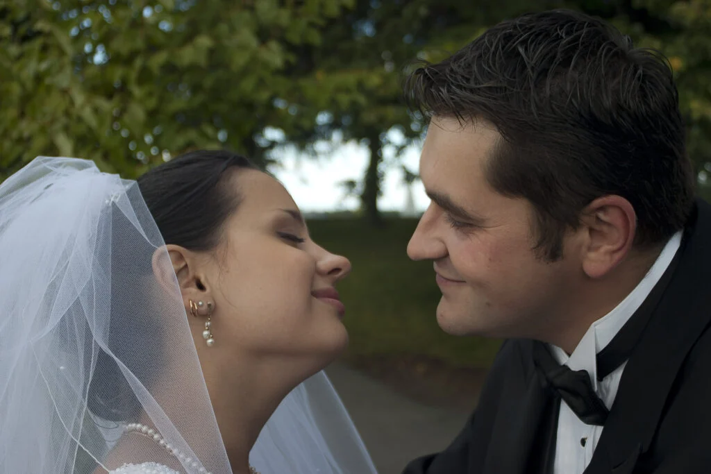 Close profile of a bride in a veil and a groom in a black bow tie smiling at each other under green trees