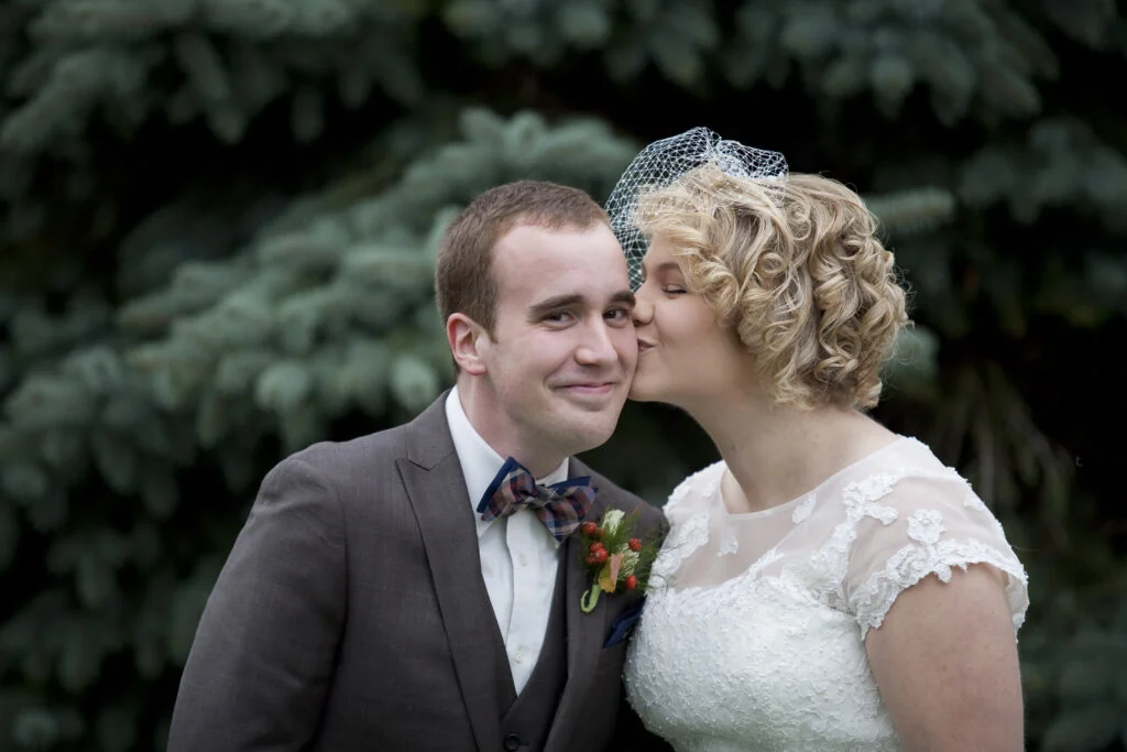A bride in a lace dress and birdcage veil kisses the groom on the cheek in front of a blue spruce
