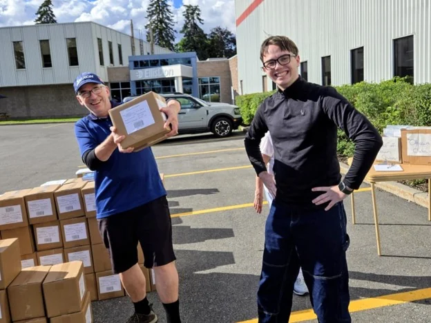 Two volunteers in a parking lot beside stacked cartons of Dictionary4Life books, one holding out a box to the camera
