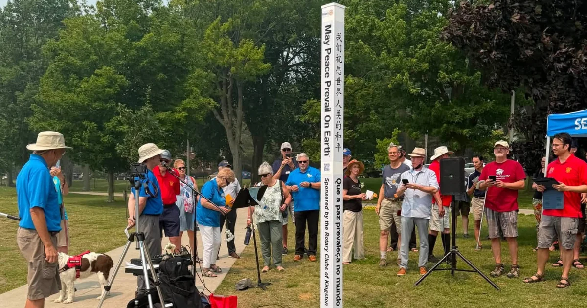A crowd in a park around a white Peace Pole reading “May Peace Prevail On Earth” in several languages, marked as sponsored by the Rotary Clubs of Kingston