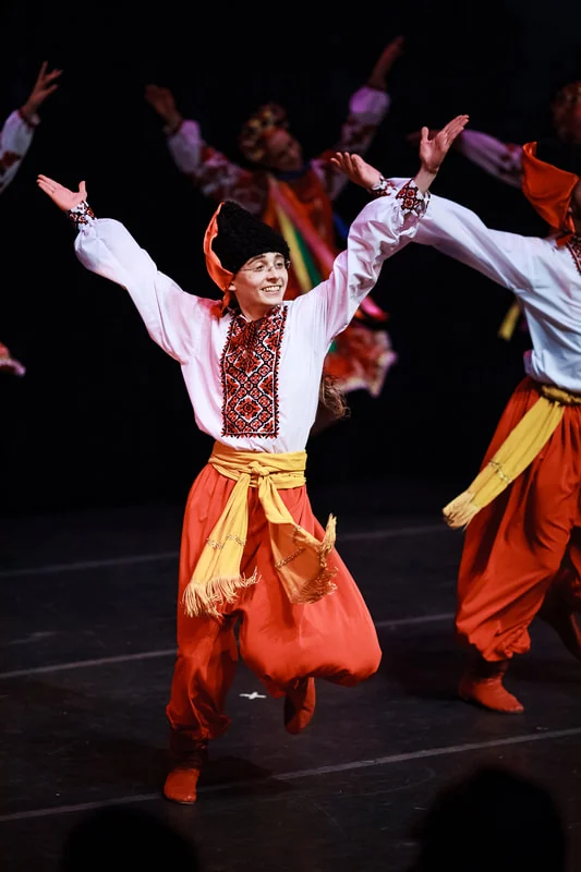 A dancer in a white embroidered shirt, orange sharovary, yellow sash and black hat, arms flung wide mid-step